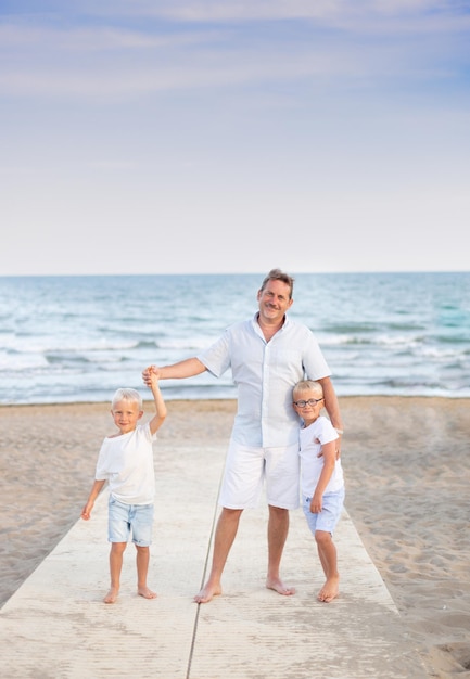 A middle aged father with 2 sons by the beach on holiday wearing light clothes and smiling