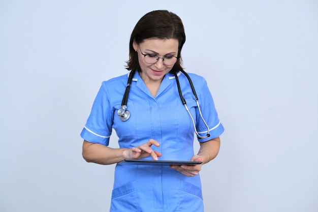 Middle-aged female medical worker with digital tablet, smiling nurse looking at tablet, reading, on gray space