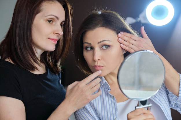 Middle-aged female patient looking in the mirror and female beautician showing on face