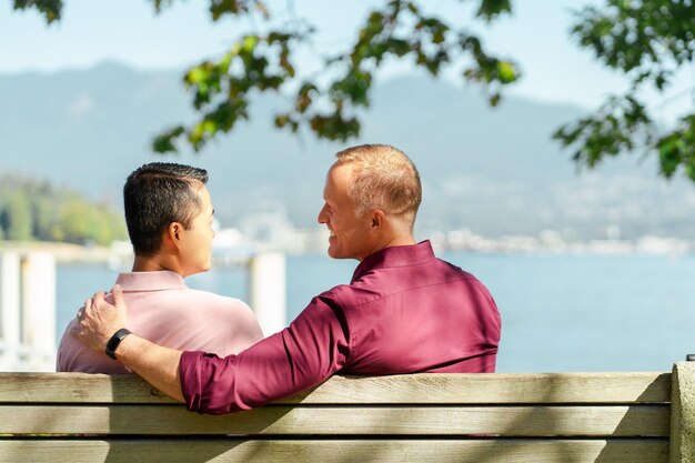 Middle aged happy homosexual couple hugging looking each other outdoors sitting on bench