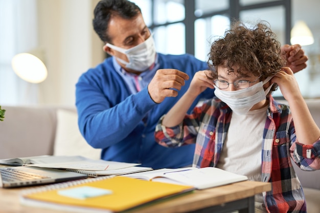 Middle aged hispanic father helps his son, school boy wearing protective mask while sitting at the desk together, using laptop and having online lesson indoors. Quarantine, homeschooling, parenthood