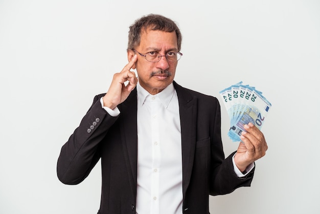 Middle aged indian business man holding bills isolated on white background pointing temple with finger, thinking, focused on a task.