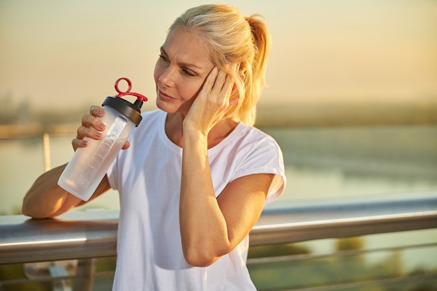 Middle-aged lady holding bottle of refreshing drink and touching her temple while suffering from migraine outdoors