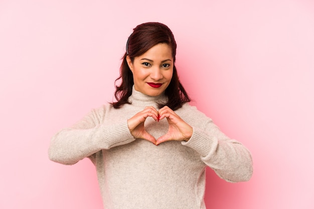 Middle aged latin woman isolated on a pink wall smiling and showing a heart shape with hands