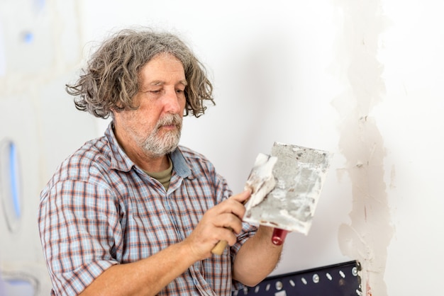 Costruttore maschio di mezza età o proprietario di una casa che ripara un muro che applica intonaco di riempimento da una piccola tavola con un raschietto mentre ripara una crepa o un'apertura.