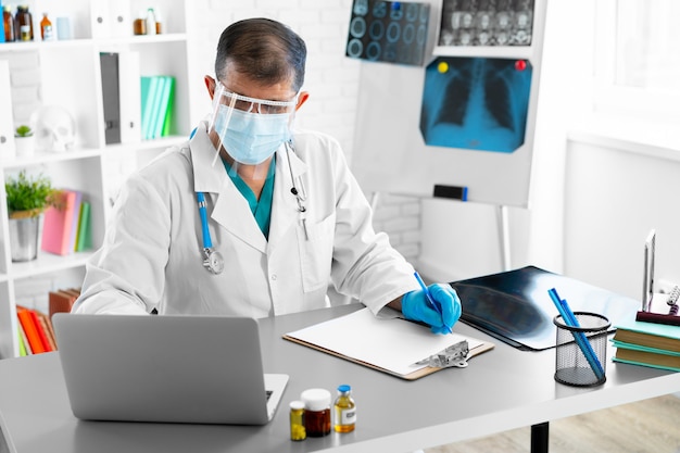 Мужчина-врач средних лет в маске и маске сидит за столом в своем офисе