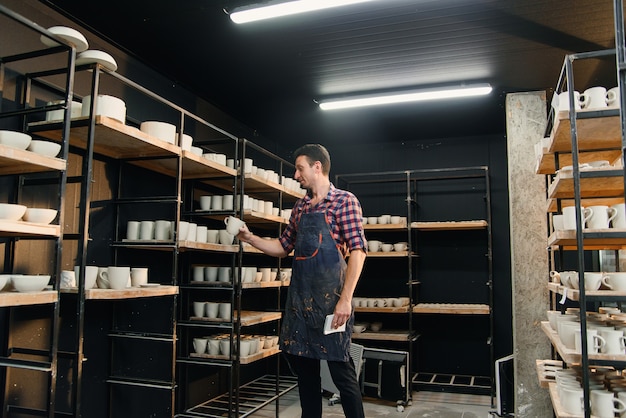 Middle aged male potter in shirt and apron counts pottery on shelves and notes their number in his notebook. Artwork and creative concept.