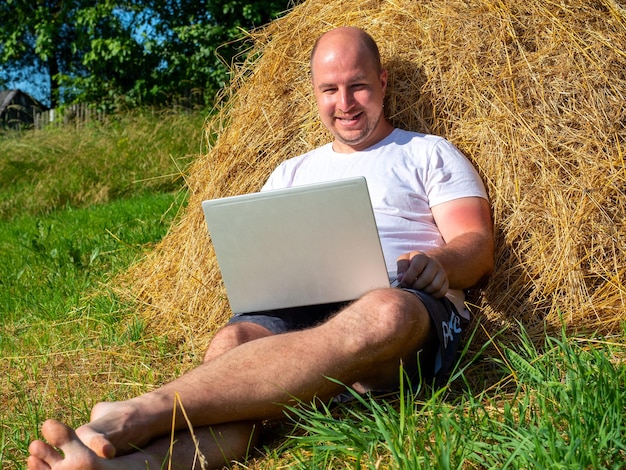 A middle-aged man, dressed in a T-shirt and shorts, is lying on a yellow haystack with a laptop in his hands. Remote work, training. Rural area. A smile on your face