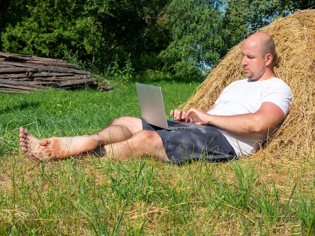 A middle-aged man dressed in a T-shirt and shorts is lying on a yellow haystack with a laptop in his hands. Remote work, training. Rural area