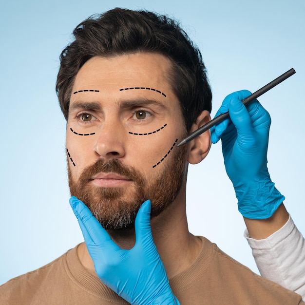 Middle aged man getting pencil marks on face skin for cosmetic surgery ready for operation blue background