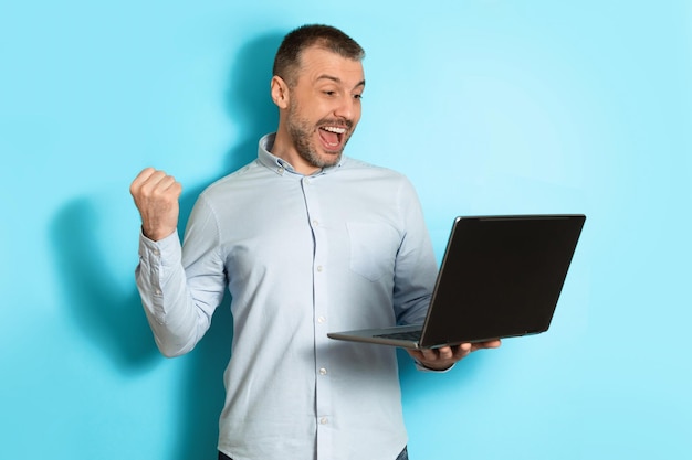 Middle aged man holding laptop computer gesturing yes blue background