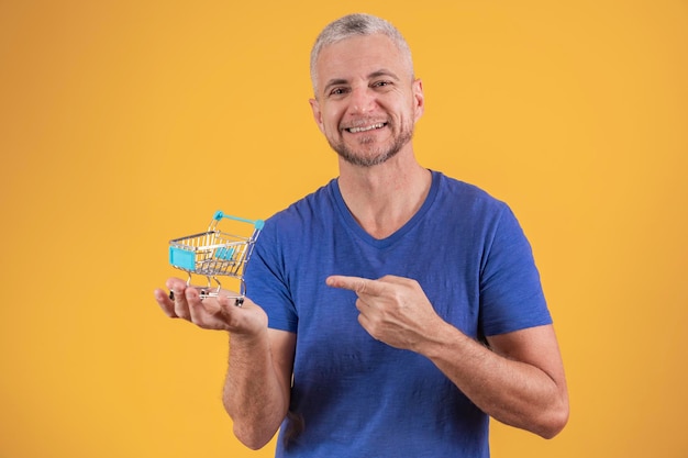 Middle aged man holding a mini shopping cart on yellow background Shopping and ecommerce concept