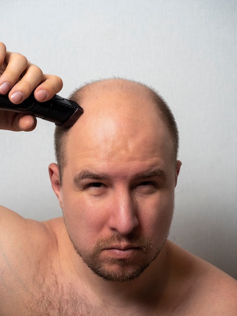 A middle-aged man looks in the mirror and shaves his head with an electric razor