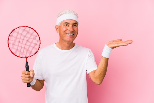 Middle aged man playing badminton isolated showing a copy space on a palm and holding another hand on waist.