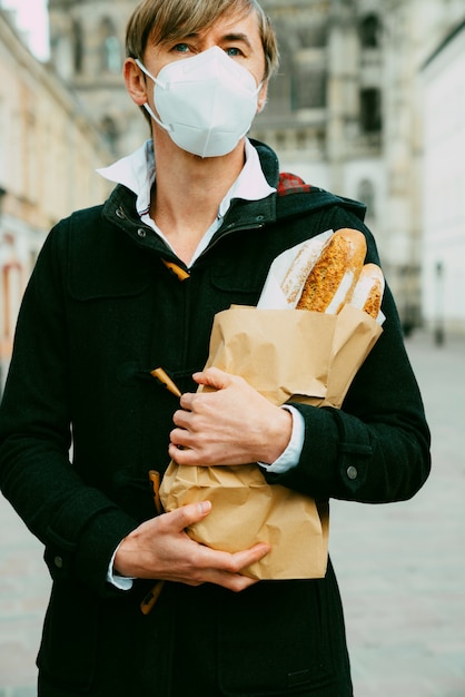 Foto uomo di mezza età per strada con pane, baguette, pagnotta durante la pandemia globale, indossando maschera, prendendo pane dal forno. cibo da asporto, spesa da forno durante il covid 19.