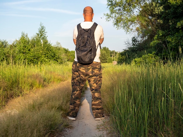 A middle-aged man with a black backpack is standing on a forest\
path at sunset. the concept of travel, walking, hiking, hiking