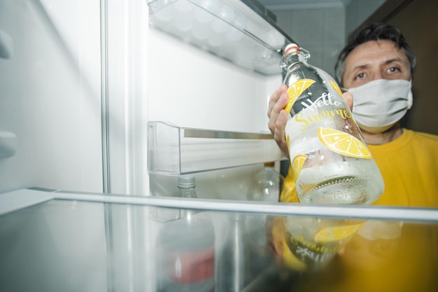 Middle-aged man with mask on his back looking at the empty fridge with one  bottle of water