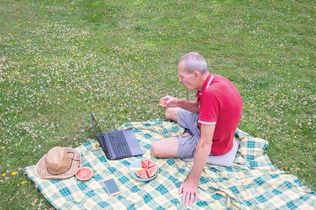 Middle aged man with mobile phone and laptop lies on a blanket in green grass