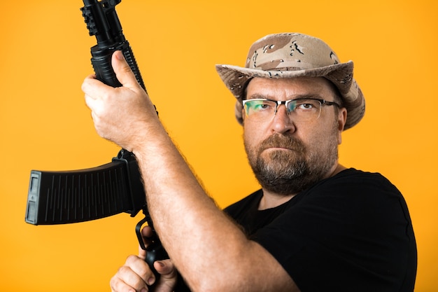 Middle aged man with rifle in black t-shirt and hat against yellow background