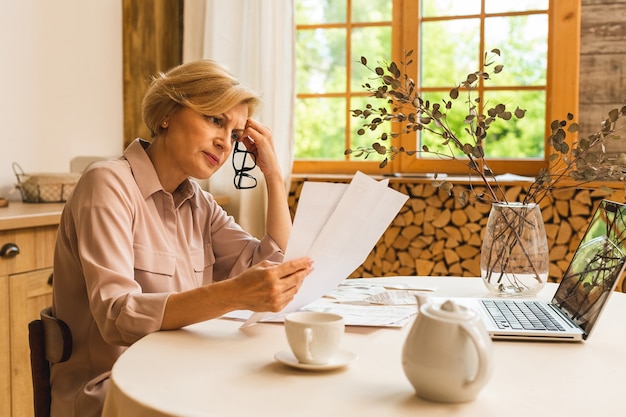 Middle aged mature senior woman holding paper bill or letter using laptop computer at home for making online payments on website, calculating financial taxes fee cost, reviewing bank account.