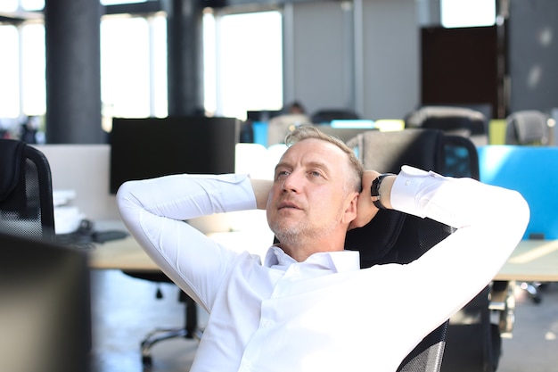 Middle aged modern businessman keeping hands behind head and smiling while sitting in the office.