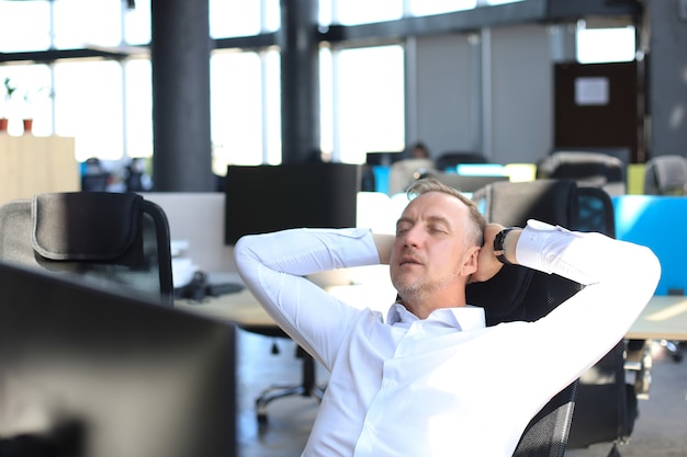 Middle aged modern businessman keeping hands behind head and smiling while sitting in the office.