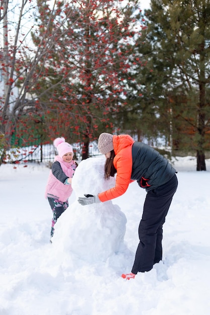 中年の母親と小さなかわいい娘が雪の上に立って、夕方に裏庭で一緒に雪だるまを作り、ナナカマドとモミの木を背景に両親が子供たちと時間を過ごす