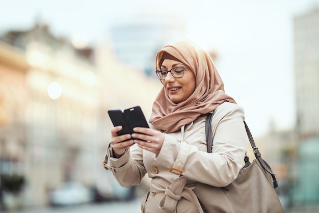 Middle aged Muslim woman wearing hijab with a happy face standing in urban environment, tipping messages on her smartphone.