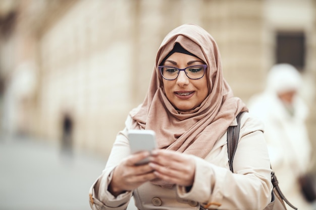 Donna musulmana di mezza età che indossa l'hijab con una faccia felice in piedi in un ambiente urbano, ribaltando i messaggi sul suo smartphone.