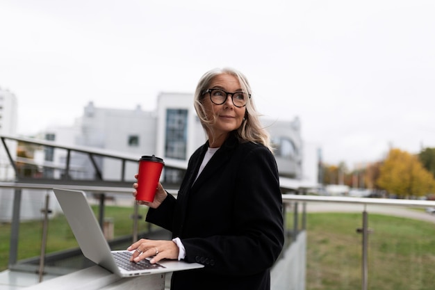 Middle aged older business woman works on a laptop and drinks coffee on the office terrace concept