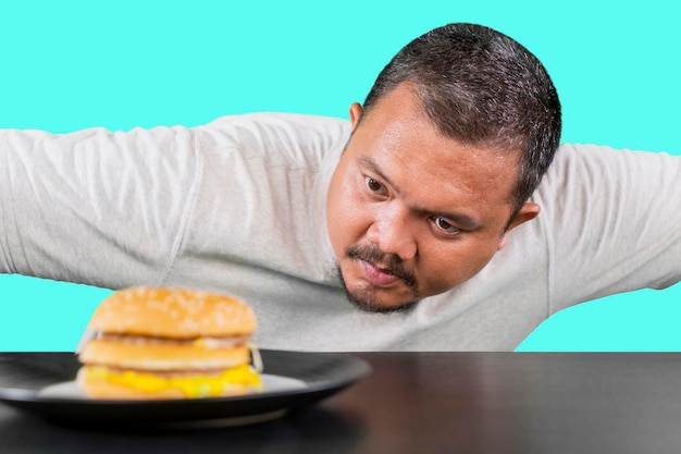 Photo middle aged overweight man looking at a cheeseburger thinking of eating it isolated on tosca background