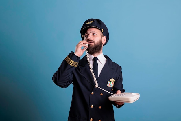 Middle aged pilot talking on landline phone, holding telephone,\
having conversation. airlane captain in professional uniform\
answering call in airport, studio medium shot