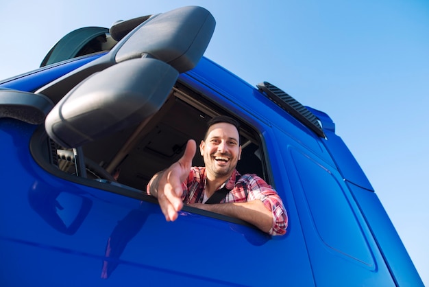 Middle aged professional truck driver sitting in his vehicle cabin and giving shaking hand to new recruits.