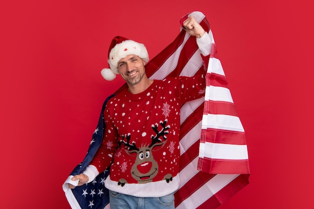 Photo middle aged santa man in christmas winter sweater on red background santa hold american flag xmas in united states usa