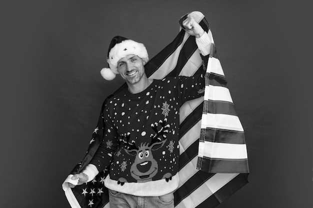 Мужчина-Санта средних лет в рождественском зимнем свитере на красном фоне Санта держит американский флаг на Рождество в США, США