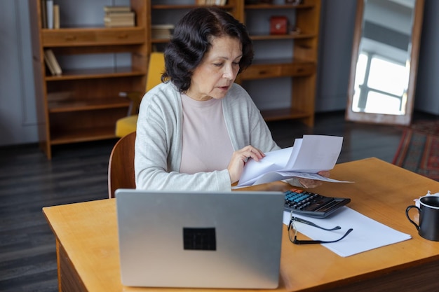 Middle aged senior woman sit with laptop and paper document pensive older mature lady reading paper