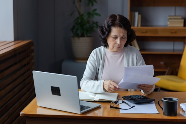 Middle aged senior woman sit with laptop and paper document pensive older mature lady reading paper