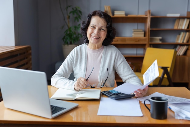 Middle aged senior woman sit with laptop and paper document Smiling older mature lady reading paper bill pay online at home managing bank finances calculating taxes planning loan debt pension payment