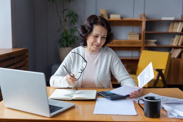 Middle aged senior woman sit with laptop and paper document Smiling older mature lady reading paper bill pay online at home managing bank finances calculating taxes planning loan debt pension payment