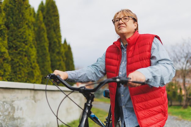 Middle aged smiling mature woman holdingt a bike with her hands\
on the grass on a green field summer or autumn country vacation and\
adventure concept