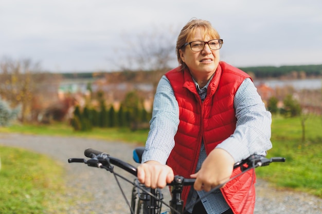 Middle aged smiling mature woman holdingt a bike with her hands\
on the grass on a green field summer or autumn country vacation and\
adventure concept