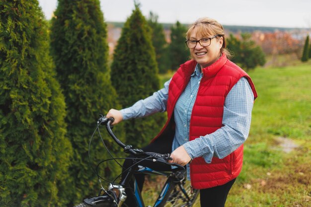 Улыбающаяся зрелая женщина средних лет держит велосипед, положив руки на траву на зеленом поле. Концепция летних или осенних загородных каникул и приключений.