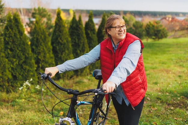 Middle aged smiling mature woman holdingt a bike with her hands\
on the grass on a green field summer or autumn country vacation and\
adventure concept