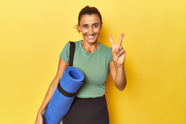 Middle aged sportswoman with yoga mat on yellow studio showing number two with fingers