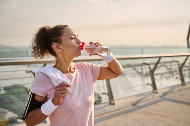 Middle-aged sporty woman in a pink T-shirt and gray leggings, with white wristbands, holds a terry towel on her shoulders and drinks water after workout standing on the city bridge at sunrise