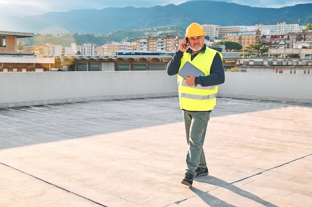 Foto supervisore di mezza età con casco e giubbotto di sicurezza che parla preoccupato sullo smartphone sul cantiere