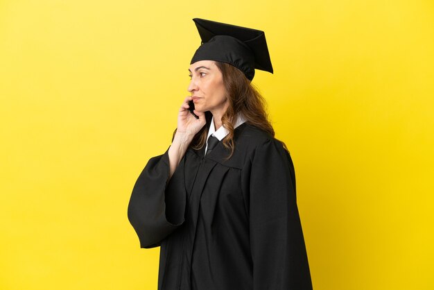 Middle aged university graduate isolated on yellow background keeping a conversation with the mobile phone with someone