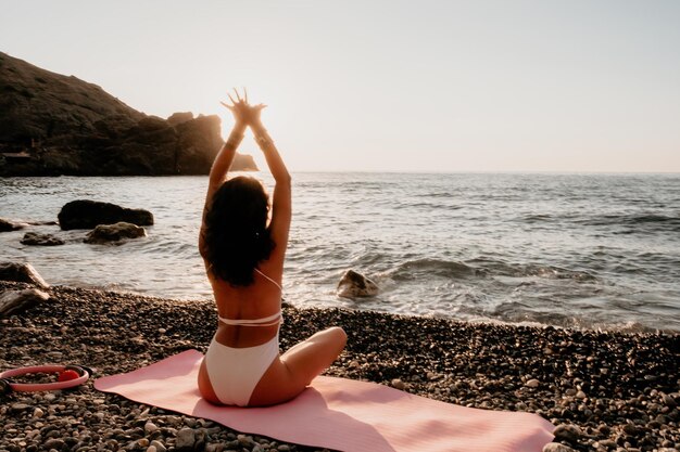 Donna di bell'aspetto di mezza età in costume da bagno bianco e bracciali in stile boho che pratica all'aperto su materassino da yoga in riva al mare su un tramonto routine di fitness yoga per donne stile di vita sano armonia e meditazione