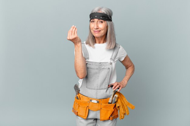 Donna di mezza età con capelli bianchi che fa un gesto capice o denaro, dicendoti di pagare e indossare abiti da lavoro e strumenti