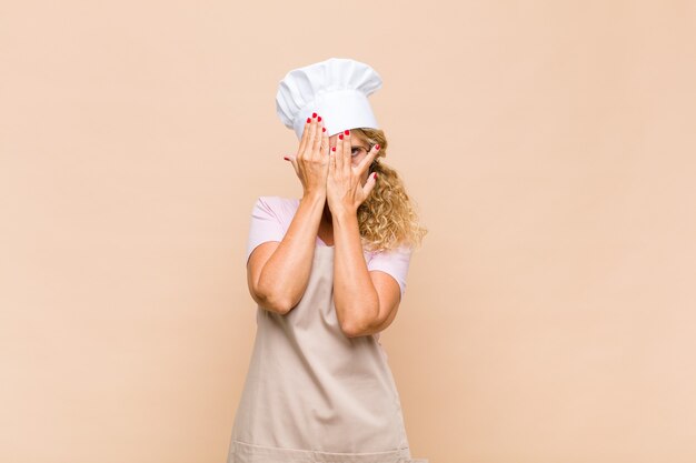 Middle aged woman baker covering face with hands, peeking between fingers with surprised expression and looking to the side